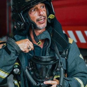 A firefighter in uniform speaking, holding a helmet indoors near fire trucks.