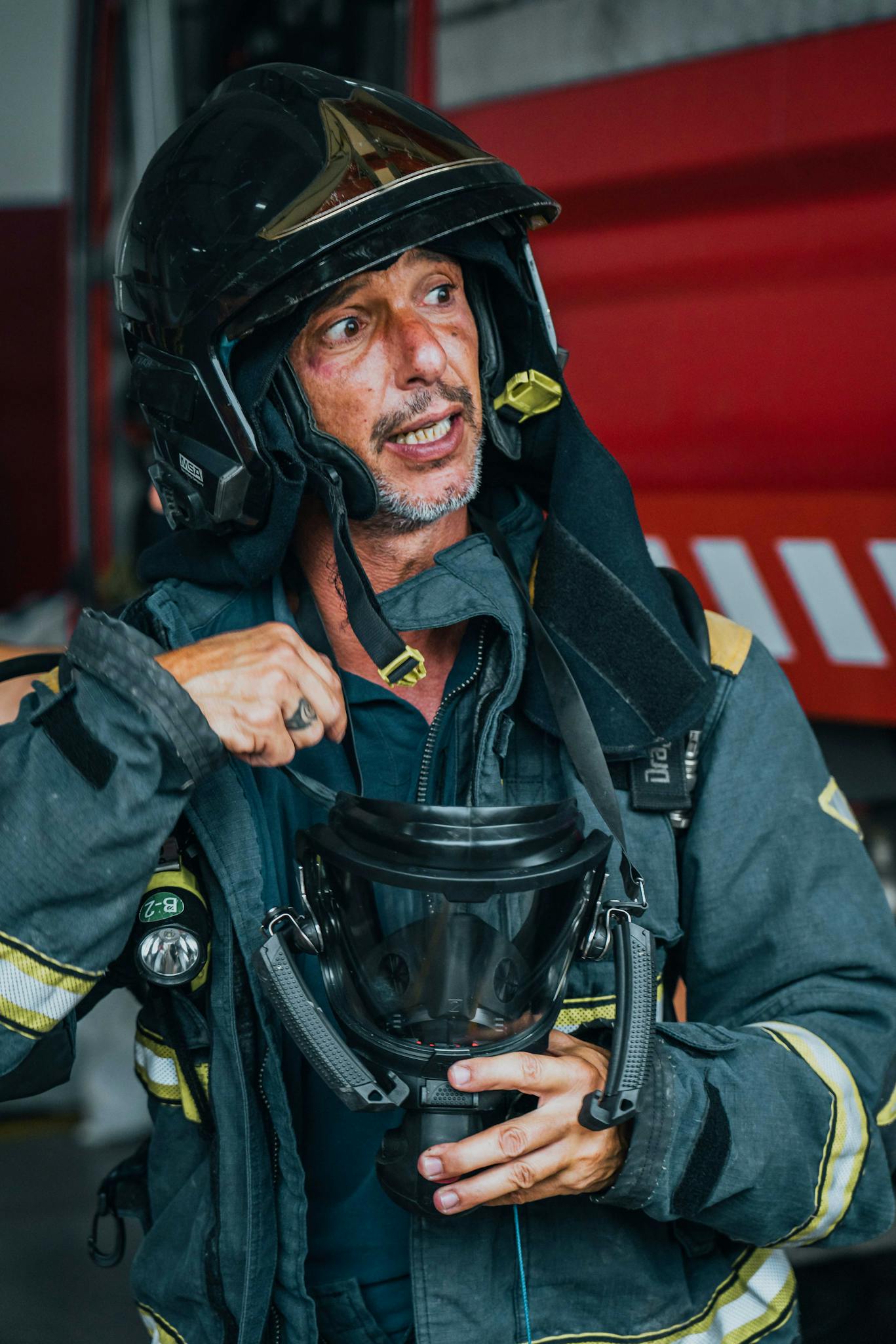 A firefighter in uniform speaking, holding a helmet indoors near fire trucks.